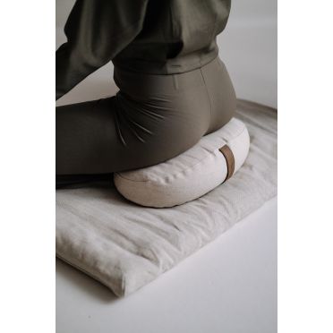 Osoba w ciemnozielonym stroju siedzi na jasnobeżowej poduszce fasolce na lnianym materacu podczas medytacji.